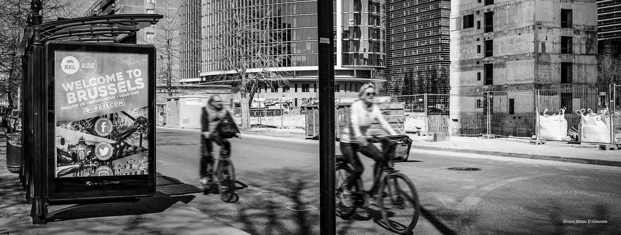 2 cyclistes de passage devant le projet Zin et les tours WTC alors qu'un panneau publicitaire nous invite à découvrir Bruxelles.